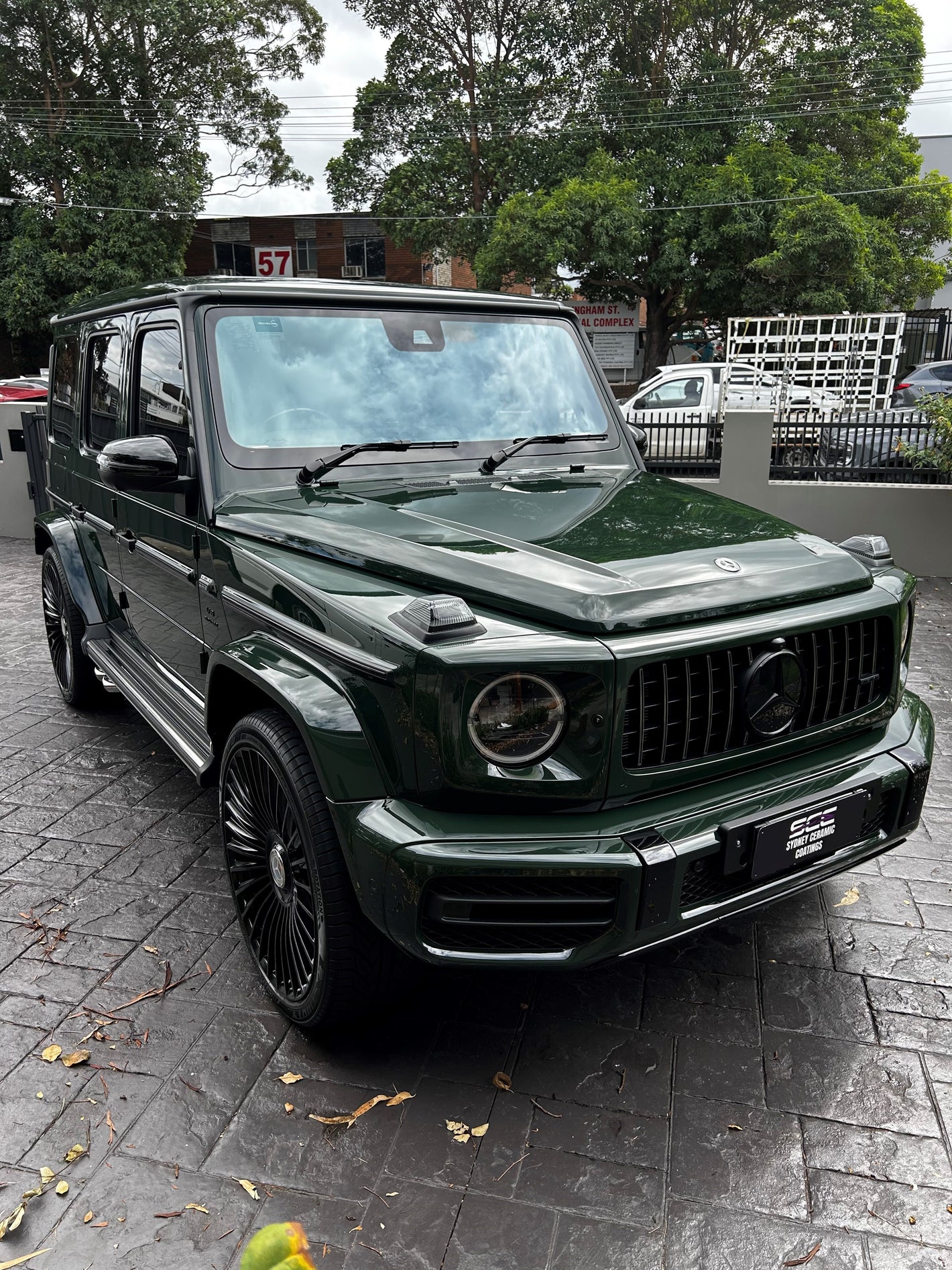Front-angle view of a dark green Mercedes-Benz G-Class SUV with blacked-out grille and rims, parked on a wet driveway near trees and buildings.